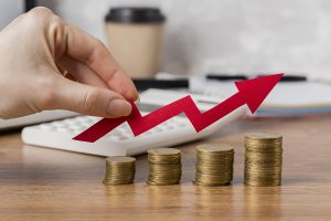 Uma mão posicionando uma seta vermelha ascendente sobre pilhas crescentes de moedas de ouro, simbolizando aumento de lucro e crescimento financeiro. Ao fundo, uma calculadora e um copo de café indicam ambiente de negócios.