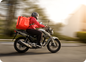 Motociclista de entrega (delivery) em alta velocidade, vestindo jaqueta vermelha e capacete, transportando uma mochila térmica grande e vermelha. A foto, com desfoque de movimento, ilustra a agilidade e eficiência da logística de entrega de bebidas.